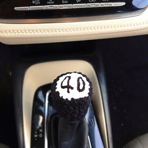 May include: A black and white crocheted gear shift knob cover featuring the number 40 stitched in black thread on a white circular top. The cover is fitted over a car gear shifter, with a textured black yarn finish and a scalloped white border.