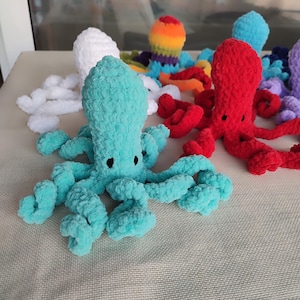 May include: A collection of colorful, handmade crochet octopuses. The octopuses are in various colors, including teal, red, white, and rainbow. Each octopus has a rounded head and eight curly tentacles. The toys are displayed on a neutral surface.