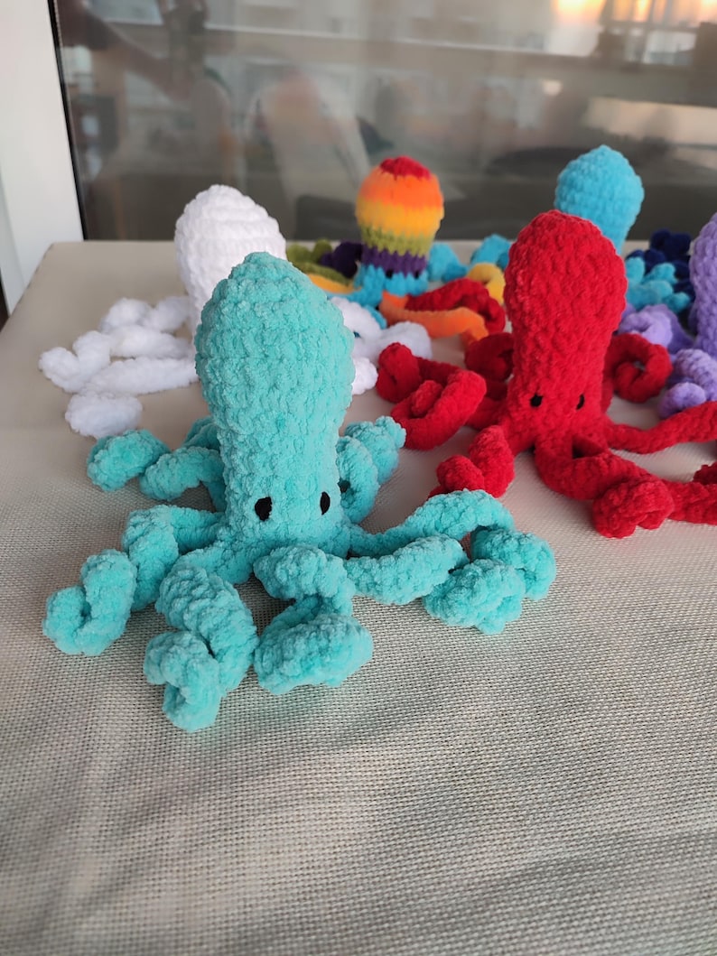 May include: A collection of colorful, handmade crochet octopuses. The octopuses are in various colors, including teal, red, white, and rainbow. Each octopus has a rounded head and eight curly tentacles. The toys are displayed on a neutral surface.