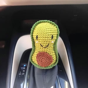 Op de afbeelding: Een gehaakt avocado auto-accessoire. De avocado heeft een lichtgroene buitenkant met een donkergroene rand en een bruine pit. Het heeft een lachend gezicht met zwarte ogen en een gebogen mond. Het accessoire is bevestigd aan een auto versnellingspook.