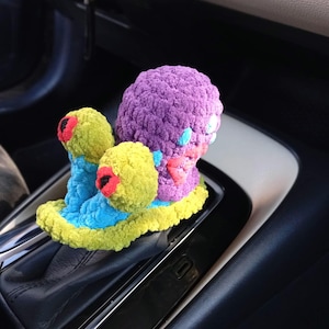 Pu&ograve; includere: Un accessorio per auto all'uncinetto fatto a mano e colorato. L'oggetto presenta una grande sfera viola con elementi gialli e blu pi&ugrave; piccoli e accenti rossi. L'oggetto &egrave; posto sul cambio di un'auto.