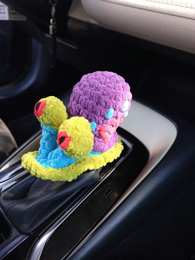 Pu&ograve; includere: Un accessorio per auto all'uncinetto fatto a mano e colorato. L'oggetto presenta una grande sfera viola con elementi gialli e blu pi&ugrave; piccoli e accenti rossi. L'oggetto &egrave; posto sul cambio di un'auto.