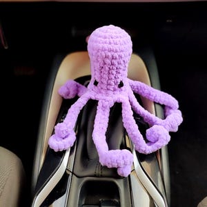Pode incluir: Um peluche de polvo em roxo claro feito de fio grosso e macio. O polvo apresenta uma cabeça arredondada, oito tentáculos enrolados e dois pequenos olhos bordados a preto. A textura do fio é visível em toda a superfície do objeto.