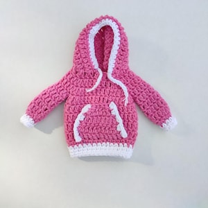 Puede incluir: Una sudadera con capucha de color rosa hecha a crochet con ribetes blancos alrededor de la capucha, las mangas y la parte inferior. La sudadera tiene un cord&oacute;n y un peque&ntilde;o bolsillo en la parte delantera.