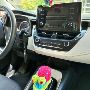 Pu&ograve; includere: Interno auto con cruscotto e cambio. Un accessorio per auto a forma di lumaca all'uncinetto rosa e blu &egrave; appeso allo specchietto retrovisore. Un'altra lumaca all'uncinetto rosa, verde e blu &egrave; sul cambio.