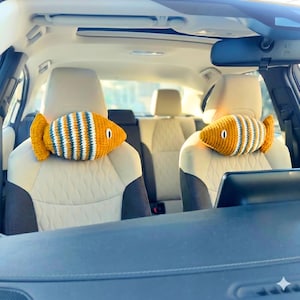 May include: Two crochet fish-shaped car headrest pillows. The pillows are yellow with blue and white stripes. The fish have a small, black eye and are placed on the headrests of a car's front seats. The car's interior is beige and black.