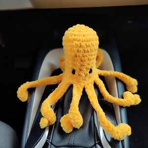 May include: A handmade, yellow crocheted octopus with black eyes. The octopus has eight long, flexible tentacles with rounded ends. The plush toy is made of soft yarn and is sitting on a dark surface.