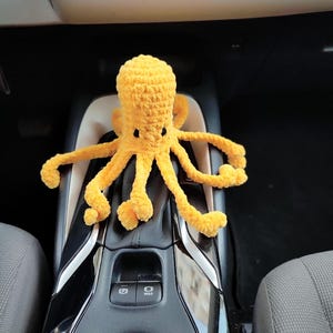 May include: A bright yellow crocheted octopus plush toy with a textured yarn surface, a rounded head, and eight long, curled tentacles. The octopus features two small, dark, oval-shaped eyes positioned on the upper front of its head.