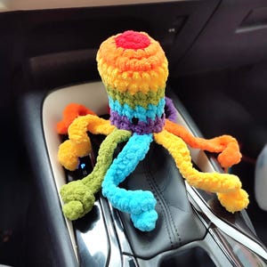 May include: A vibrant, crocheted octopus toy. The octopus has a rainbow-coloured head and tentacles in shades of orange, yellow, green, blue, and purple. The plush toy is soft and appears to be handmade.