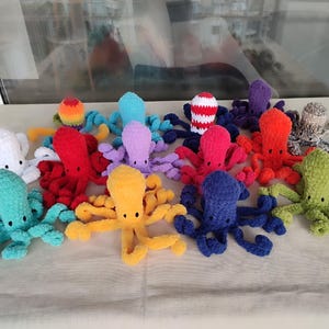 May include: A collection of colorful, handmade crochet octopuses. Each octopus features a round head and eight long tentacles. The octopuses are in various colors, including white, red, yellow, blue, green, and purple. The octopuses are displayed on a light-colored surface.