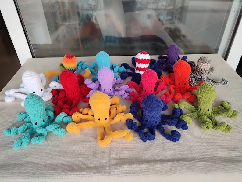 May include: A collection of colorful, handmade crochet octopuses. Each octopus features a round head and eight long tentacles. The octopuses are in various colors, including white, red, yellow, blue, green, and purple. The octopuses are displayed on a light-colored surface.