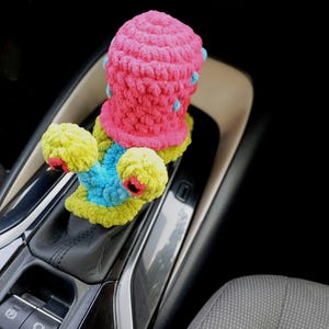 Pu&ograve; includere: Accessorio per auto all'uncinetto fatto a mano e colorato. L'oggetto presenta una parte superiore rosa vibrante con dettagli blu, una base gialla e dettagli blu e rossi. L'accessorio &egrave; progettato per adattarsi alla leva del cambio di un'auto.