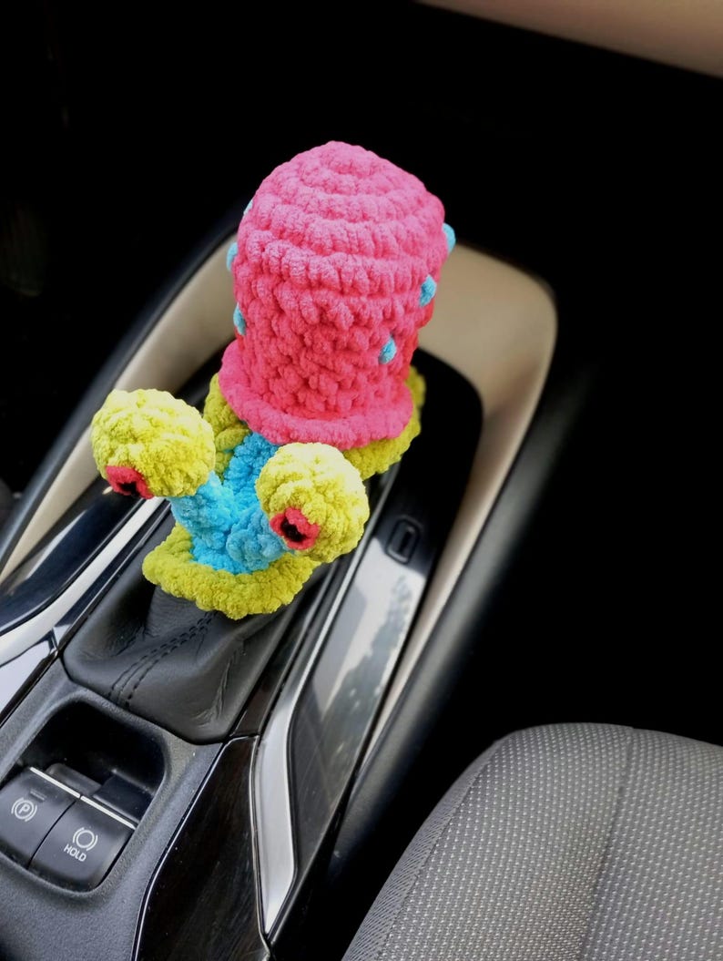 Pu&ograve; includere: Accessorio per auto all'uncinetto fatto a mano e colorato. L'oggetto presenta una parte superiore rosa vibrante con dettagli blu, una base gialla e dettagli blu e rossi. L'accessorio &egrave; progettato per adattarsi alla leva del cambio di un'auto.