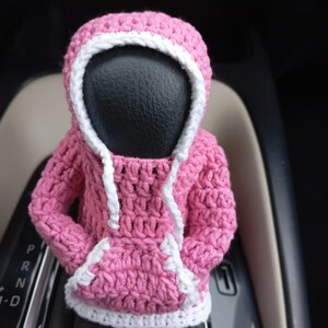 Puede incluir: Una sudadera con capucha de color rosa hecha a crochet con ribetes blancos, dise&ntilde;ada para ajustarse a la palanca de cambios de un coche.