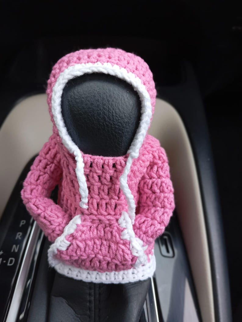 Puede incluir: Una sudadera con capucha de color rosa hecha a crochet con ribetes blancos, dise&ntilde;ada para ajustarse a la palanca de cambios de un coche.