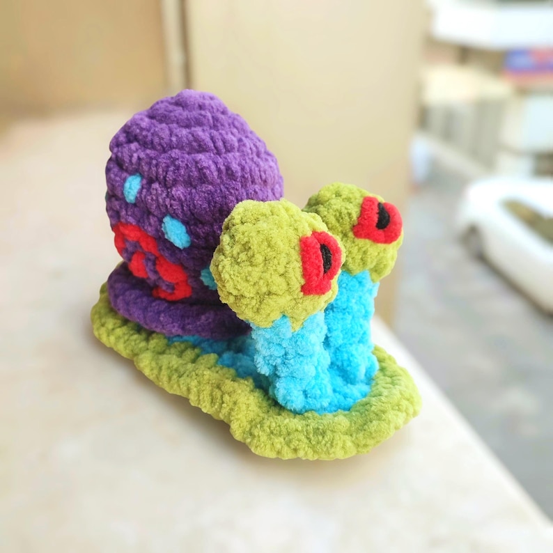Pu&ograve; includere: Un giocattolo a forma di lumaca fatto a mano, in colori vivaci. Il guscio della lumaca &egrave; viola strutturato, con dettagli blu e rossi. Il suo corpo &egrave; blu brillante e gli occhi sono verdi con dettagli rossi. La lumaca poggia su una base verde.