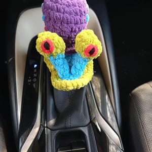 Pu&ograve; includere: Un accessorio per auto fatto a mano, all'uncinetto con colori vivaci. Presenta una testa viola, braccia gialle con dettagli rossi e neri e tocchi di blu. Progettato per adattarsi alla leva del cambio di un'auto.