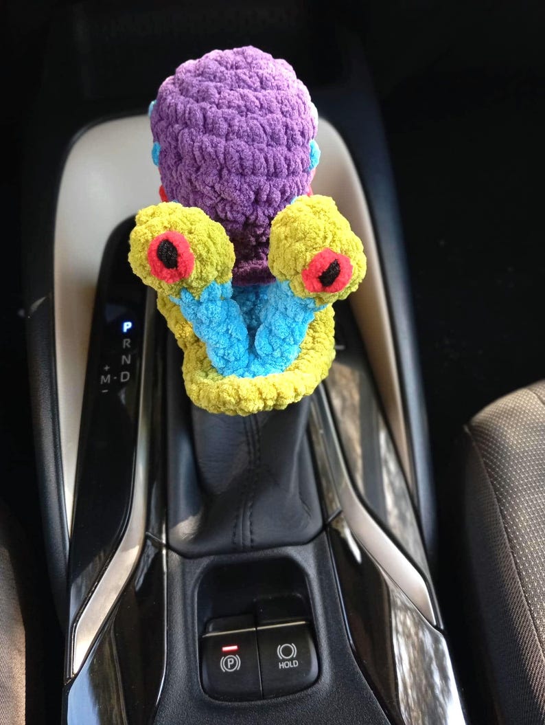 Pu&ograve; includere: Un accessorio per auto fatto a mano, all'uncinetto con colori vivaci. Presenta una testa viola, braccia gialle con dettagli rossi e neri e tocchi di blu. Progettato per adattarsi alla leva del cambio di un'auto.