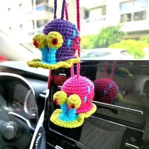 Pu&ograve; includere: Due ciondoli per auto a forma di lumaca fatti a mano all'uncinetto. Uno &egrave; viola con dettagli blu e rossi, l'altro &egrave; rosa con dettagli blu. Entrambi hanno occhi gialli e basi blu e gialle. Sono appesi a cordoncini viola e rosa.