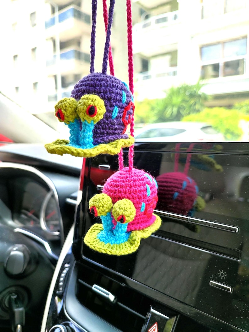 Pu&ograve; includere: Due ciondoli per auto a forma di lumaca fatti a mano all'uncinetto. Uno &egrave; viola con dettagli blu e rossi, l'altro &egrave; rosa con dettagli blu. Entrambi hanno occhi gialli e basi blu e gialle. Sono appesi a cordoncini viola e rosa.