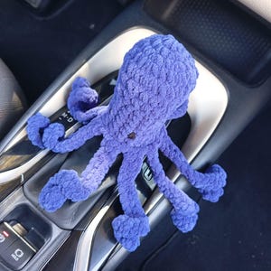 May include: A handmade, plush, purple octopus toy with eight tentacles. The octopus is made of a soft, textured material and has a small, dark button for an eye. The toy is resting on a car's center console.
