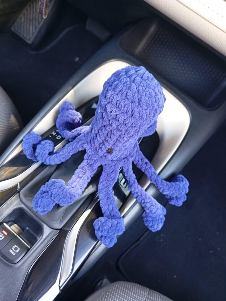 May include: A handmade, plush, purple octopus toy with eight tentacles. The octopus is made of a soft, textured material and has a small, dark button for an eye. The toy is resting on a car's center console.