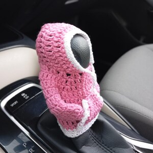 Puede incluir: Cubierta de palanca de cambios de coche de ganchillo rosa con ribete blanco. La cubierta tiene forma de figura con capucha y cara negra.