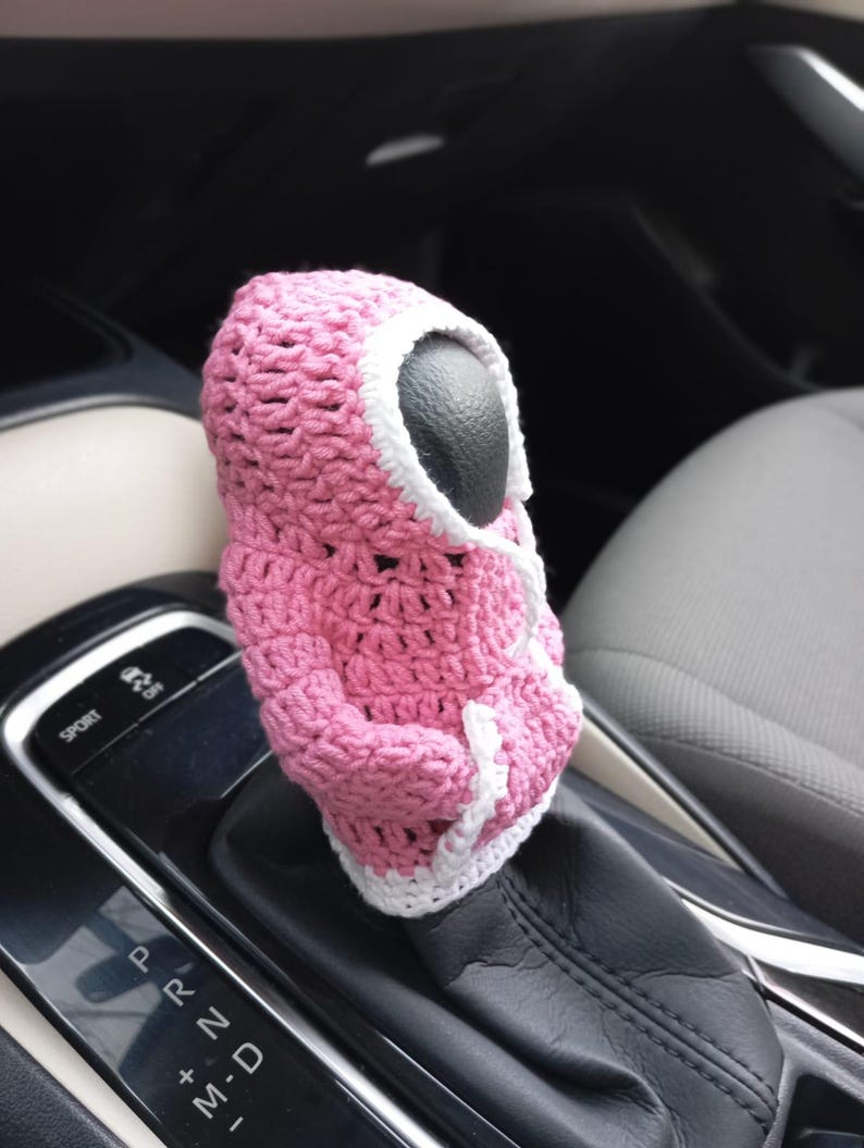 Puede incluir: Cubierta de palanca de cambios de coche de ganchillo rosa con ribete blanco. La cubierta tiene forma de figura con capucha y cara negra.