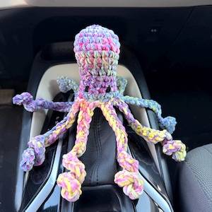 Könnte beinhalten: Ein handgefertigtes Häkel-Oktopus-Spielzeug aus mehrfarbigem Garn in Rosa, Gelb, Blau und Violett. Das Plüschtier hat einen abgerundeten Kopf, acht lange, geringelte Tentakel und zwei kleine, dunkelbraun gestickte Augen auf der Vorderseite.
