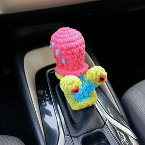 Pu&ograve; includere: Una copertura per il cambio dell'auto all'uncinetto, fatta a mano e colorata. La copertura presenta una parte superiore rosa con accenti blu e una base verde e blu con dettagli rossi e neri. Progettata per adattarsi al cambio dell'auto.