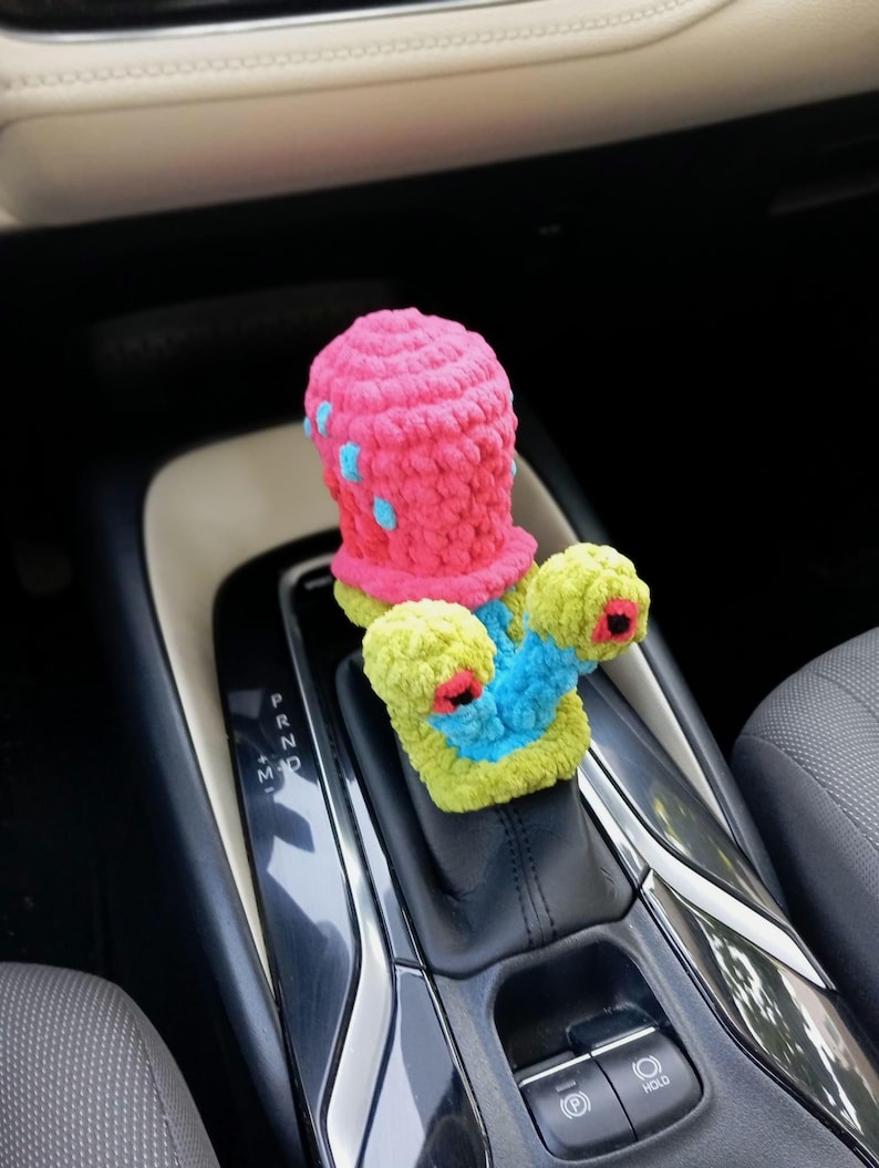 Pu&ograve; includere: Una copertura per il cambio dell'auto all'uncinetto, fatta a mano e colorata. La copertura presenta una parte superiore rosa con accenti blu e una base verde e blu con dettagli rossi e neri. Progettata per adattarsi al cambio dell'auto.