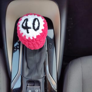 Peut inclure: Un couvre-pommeau de levier de vitesses crocheté à la main, de couleur rouge, avec le chiffre "40" en noir entouré d'une bordure blanche. Il est installé sur le levier de vitesses d'une voiture, ajoutant une touche personnalisée à l'intérieur.