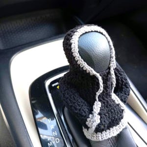 Op de afbeelding: Een zwarte en grijze gehaakte auto versnellingspookhoes. De hoes is gevormd als een capuchontrui met een rits.