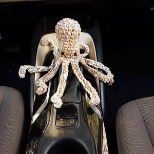 May include: A handmade crochet octopus in shades of beige and brown, resting on a car's console. The octopus has eight long, textured tentacles and small black eyes. The car's interior is visible in the background.