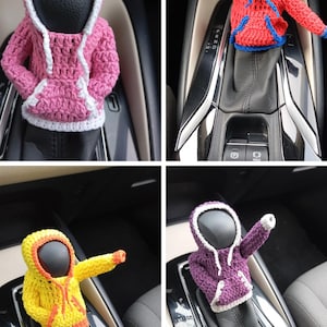 Puede incluir: Se muestran cuatro figuras con capucha de ganchillo en diferentes colores, rosa, rojo, amarillo y morado, en la palanca de cambios de un coche. Las figuras llevan sudaderas con capucha con ribetes blancos.
