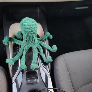 May include: A handmade, green crocheted octopus plush toy. The octopus has eight long, curled tentacles and a textured body. The plush is sitting on a car's center console, with the gear shift visible.