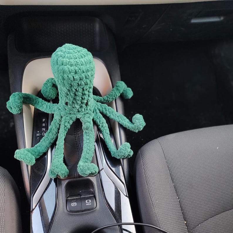 May include: A handmade, green crocheted octopus plush toy. The octopus has eight long, curled tentacles and a textured body. The plush is sitting on a car's center console, with the gear shift visible.