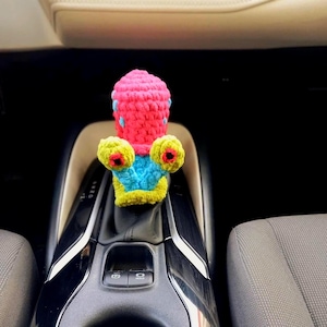 Puede incluir: Una funda de pomo de cambio de ganchillo hecha a mano y colorida. La funda es principalmente rosa con detalles azules y presenta dos grandes formas de ojos amarillos y azules. Diseñada para la palanca de cambios de un coche.
