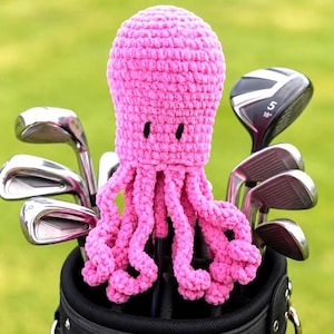 May include: A bright pink crocheted octopus golf club cover with two small black embroidered eyes, featuring eight dangling tentacles, positioned inside a black golf bag surrounded by several metal golf clubs with silver heads.
