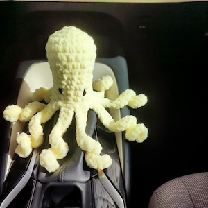 Peut inclure: Une pieuvre au crochet de couleur jaune pâle. Le jouet en peluche a une tête arrondie, des yeux noirs et huit tentacules bouclés. La pieuvre est assise sur un siège de voiture.
