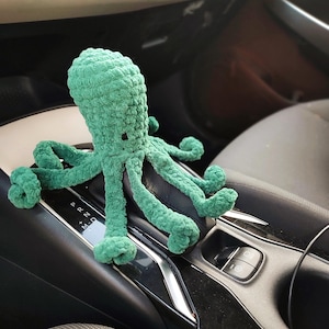 May include: A green crocheted octopus toy with eight tentacles. The plush toy is positioned on a car's center console, with the gear shift and other controls in the background. The octopus has a textured, handmade appearance.