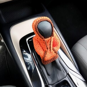 May include: Orange and brown crocheted hooded figure covering a car's gear shift lever. The gear shift lever has the letters "M, D, N, P, S" printed on it.