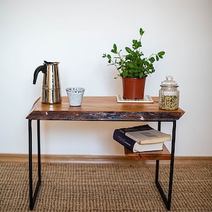 Op de afbeelding: Een houten salontafel met een zwart metalen frame en een lager plankje. De tafel heeft een live edge-ontwerp en is versierd met een plant in een pot, een koffiekan, een kopje en een pot gedroogde kruiden.