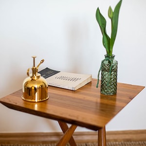 Könnte beinhalten: Ein Holz-Beistelltisch mit einer Messing-Gießkanne, einem Buch mit dem Titel "COLLATERAL" und einer grünen Glasvase mit einer einzelnen weißen Tulpe.