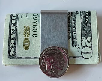 May include: A stainless steel money clip holding a stack of twenty-dollar bills and a buffalo nickel. The bills are green and white, and the nickel is silver. The money clip is rectangular and has a brushed metal finish.