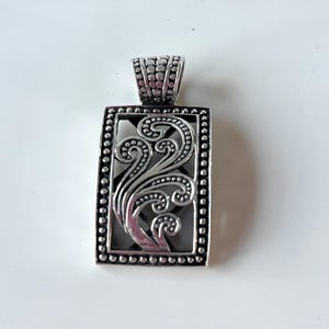 May include: Silver-toned rectangular pendant with an ornate, swirling floral design. The pendant has a beaded border and a textured bail for a chain. The intricate details and polished finish give it a classic, elegant look.