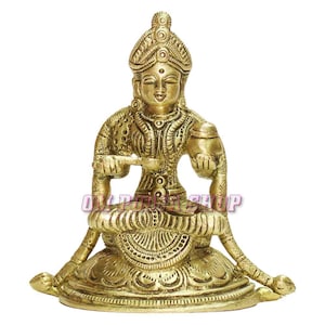May include: A gold-colored brass statue of a seated Hindu goddess. She is holding a spoon in one hand and a pot in the other. She is wearing a crown and ornate clothing. The statue is sitting on a lotus base.