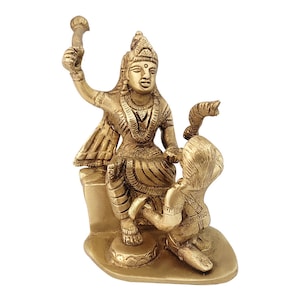 May include: A gold-colored brass statue of a Hindu goddess sitting on a throne with a snake and a smaller figure kneeling before her.
