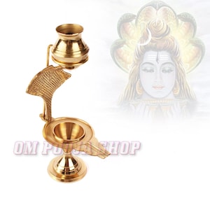 May include: A gold brass diya with a cobra hood design and a small pot on top. The diya is on a white background with a blurred image of Shiva in the background.