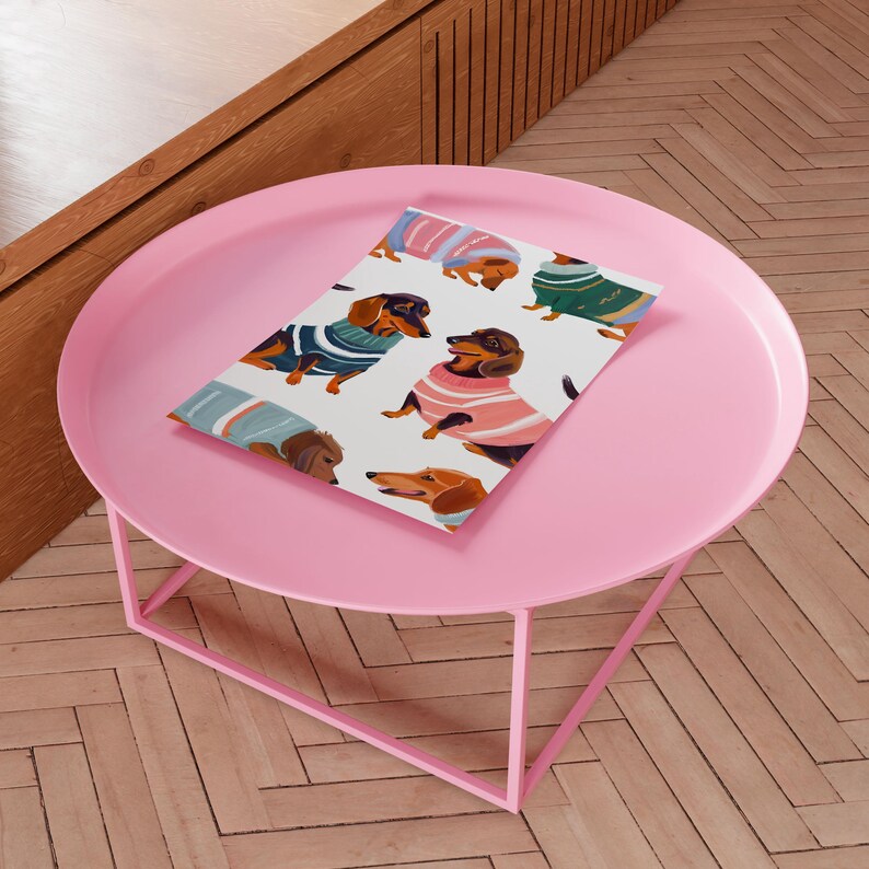 May include: A pink round table with a white background featuring a repeating pattern of dachshund dogs wearing colorful sweaters. The table has a square metal base.